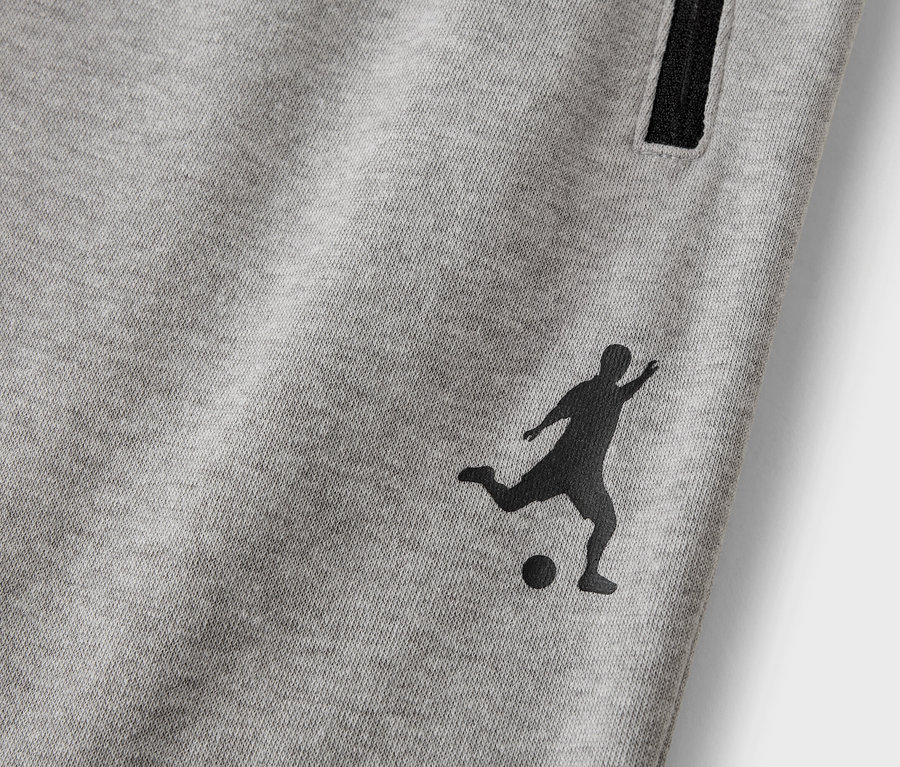 Detail šedých teplákov s čiernou siluetou futbalistu a vreckom na zips.