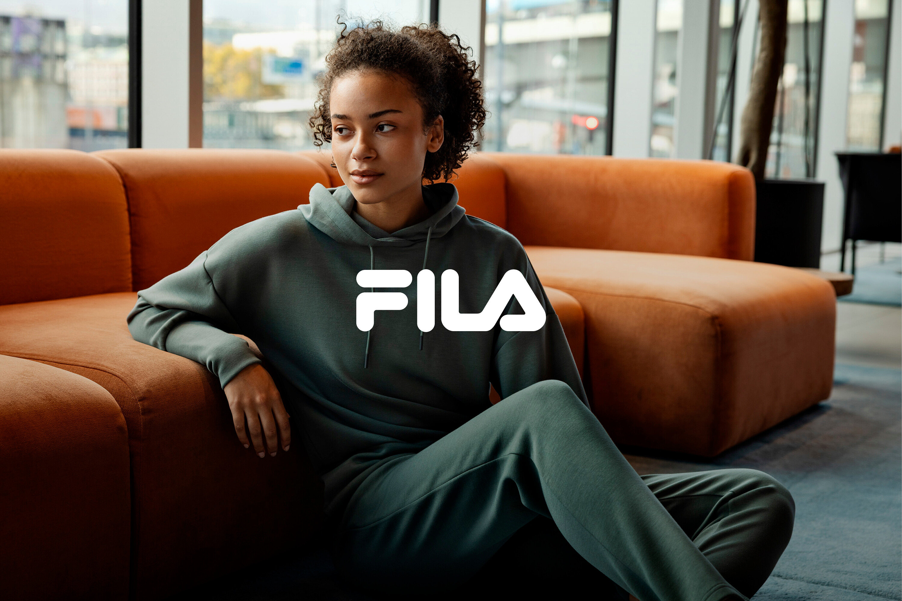 Žena sedí na zemi, opretá o oranžovú pohovku, oblečená v zelenej teplákovej súprave Fila.