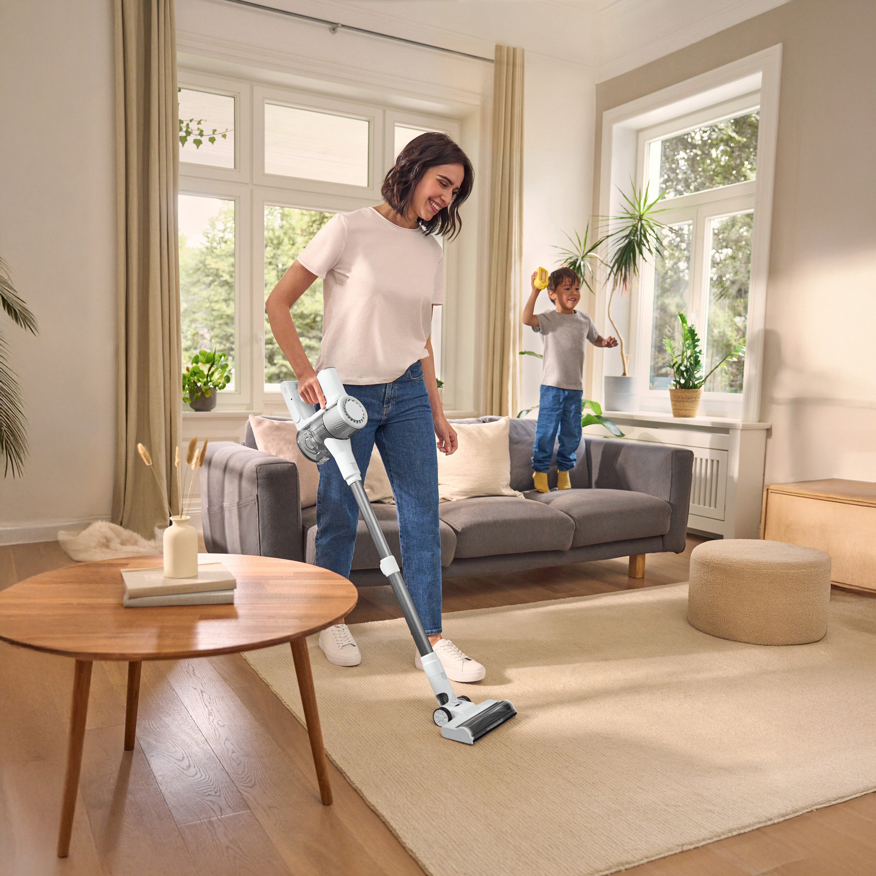Žena vysáva koberec akumulátorovým vysávačom, dieťa stojí na pohovke. Okrúhly stôl vľavo, okno v pozadí.
