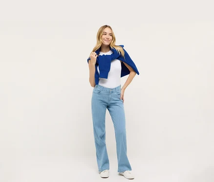 Žena pózuje v bielom tričku a modrých džínsoch Angels Denim »Liz«, Wide Leg, s modrým svetrom prehodeným cez ramená.