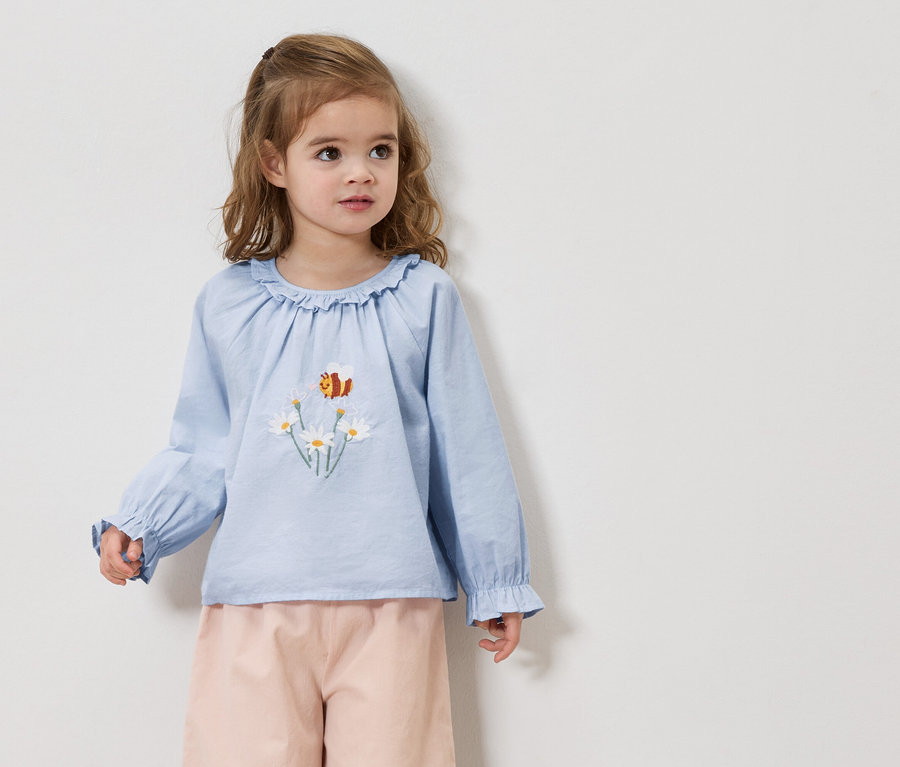 Dievča s blond vlasmi má na sebe svetlomodrú blúzku s výšivkou sedmokrások a včiel a ružové nohavice.