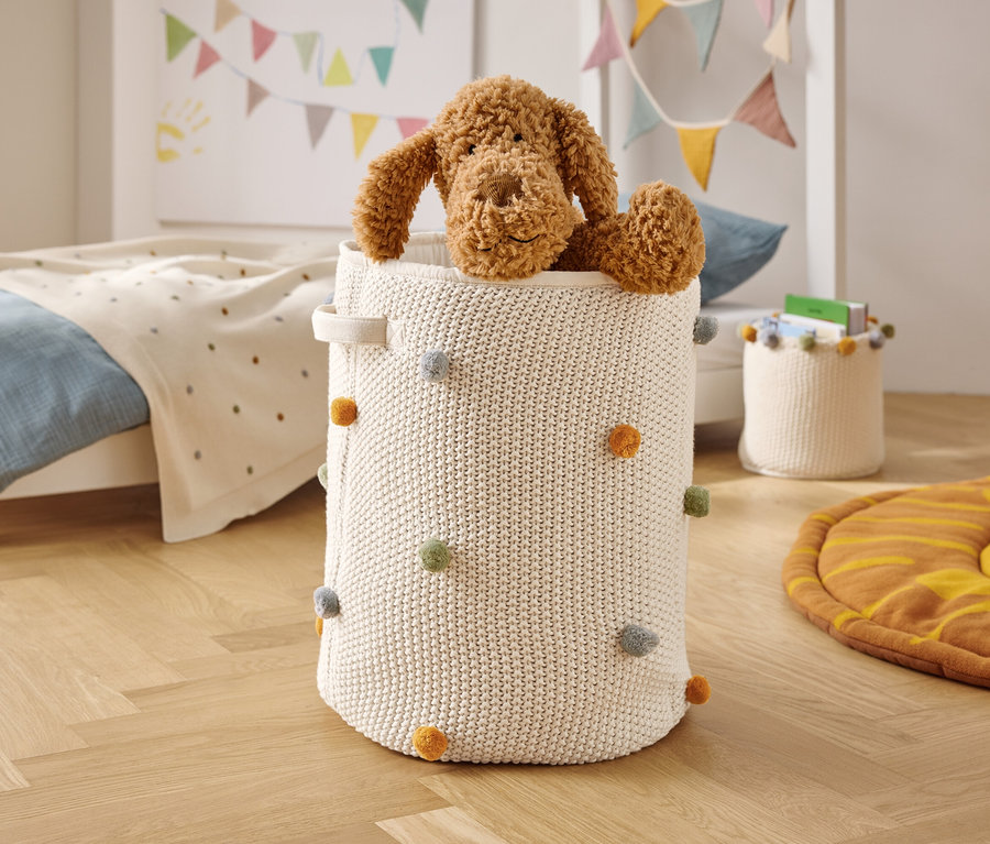 Hnedý plyšový pes sedí v bielom, pletenom koši s farebnými brmbolcami.