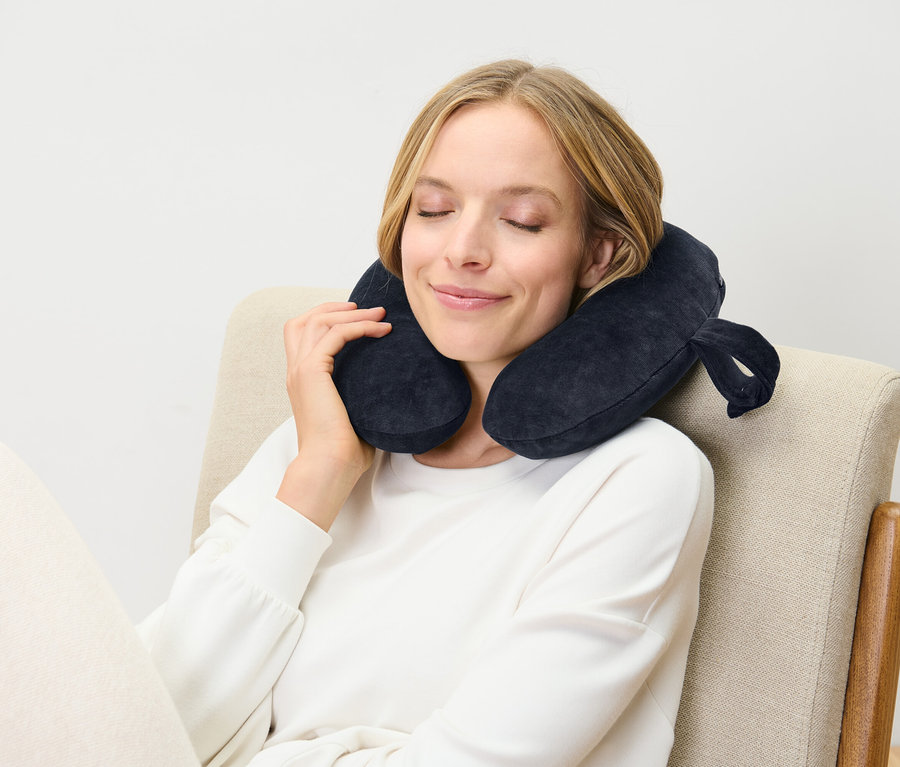 Žena sedí so zatvorenými očami na stoličke a má na sebe tmavomodrý cestovný vankúš.