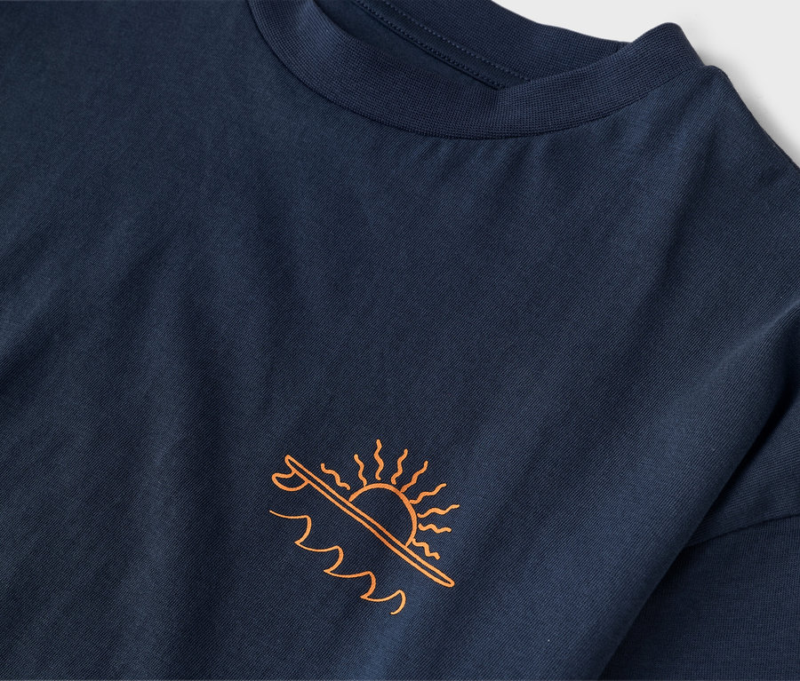 Detailný záber na tmavomodré tričko s oranžovou potlačou slnka nad surfovou doskou a vlnami.