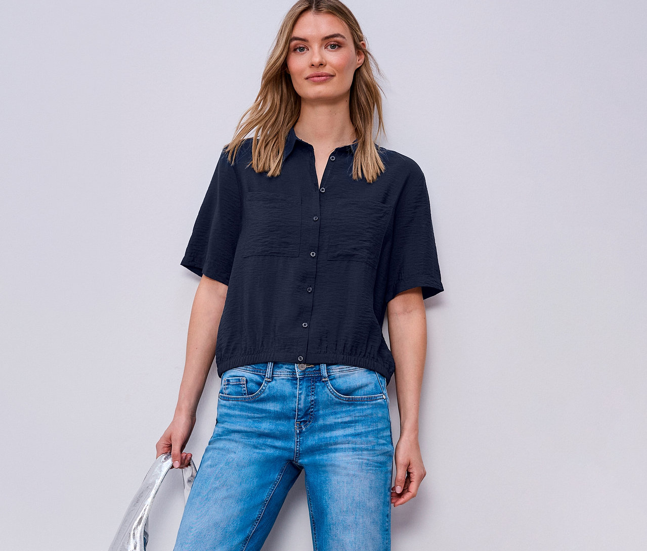 Žena má na sebe džínsy a košeľovú blúzku Street One. Drží striebornú tašku.