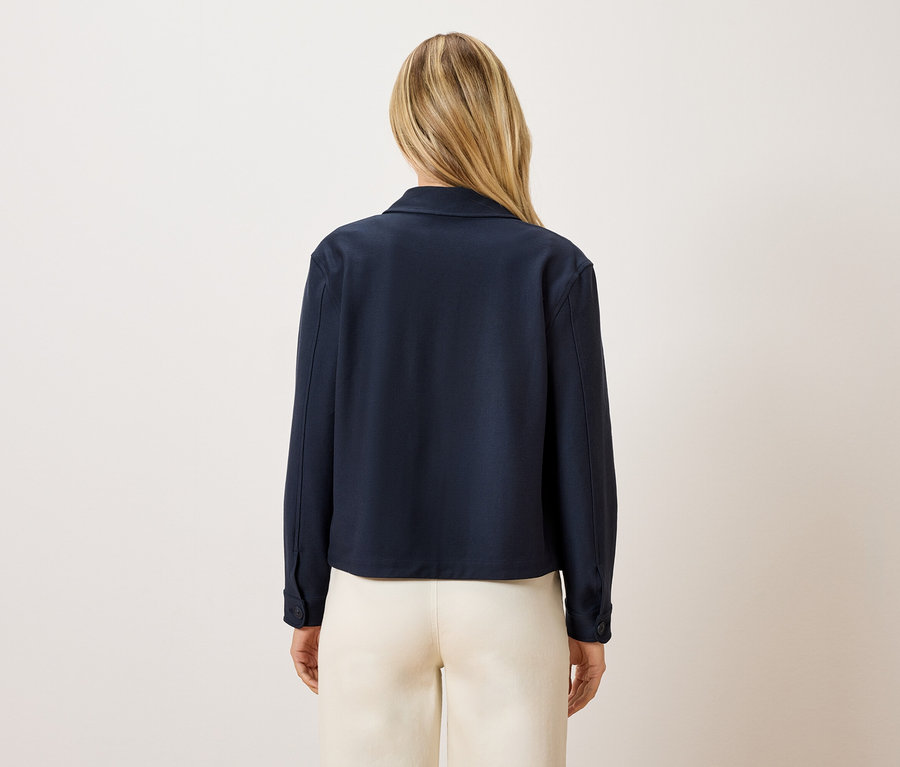 Pohľad zozadu na ženu s blond vlasmi v tmavomodrej bunde a krémových nohaviciach.