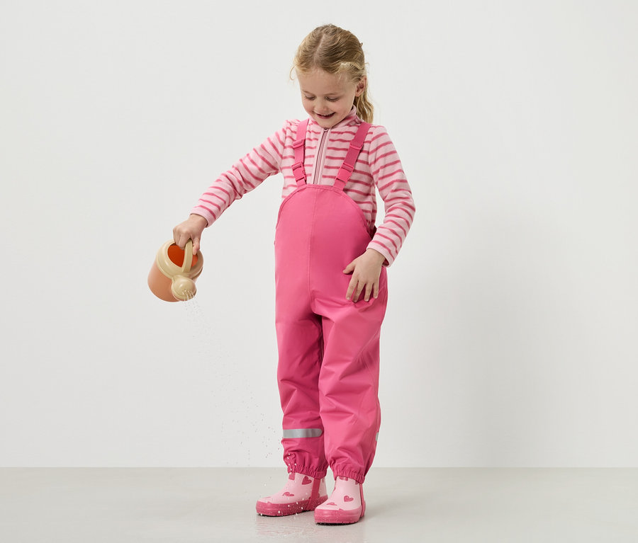 Dievča polieva vodu krhlou, oblečené v ružových záhradníkoch a topánkach.