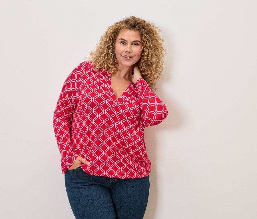 Žena s kučeravými vlasmi v červeno-bielej blúzke a džínsoch pózuje s rukou vo vrecku.