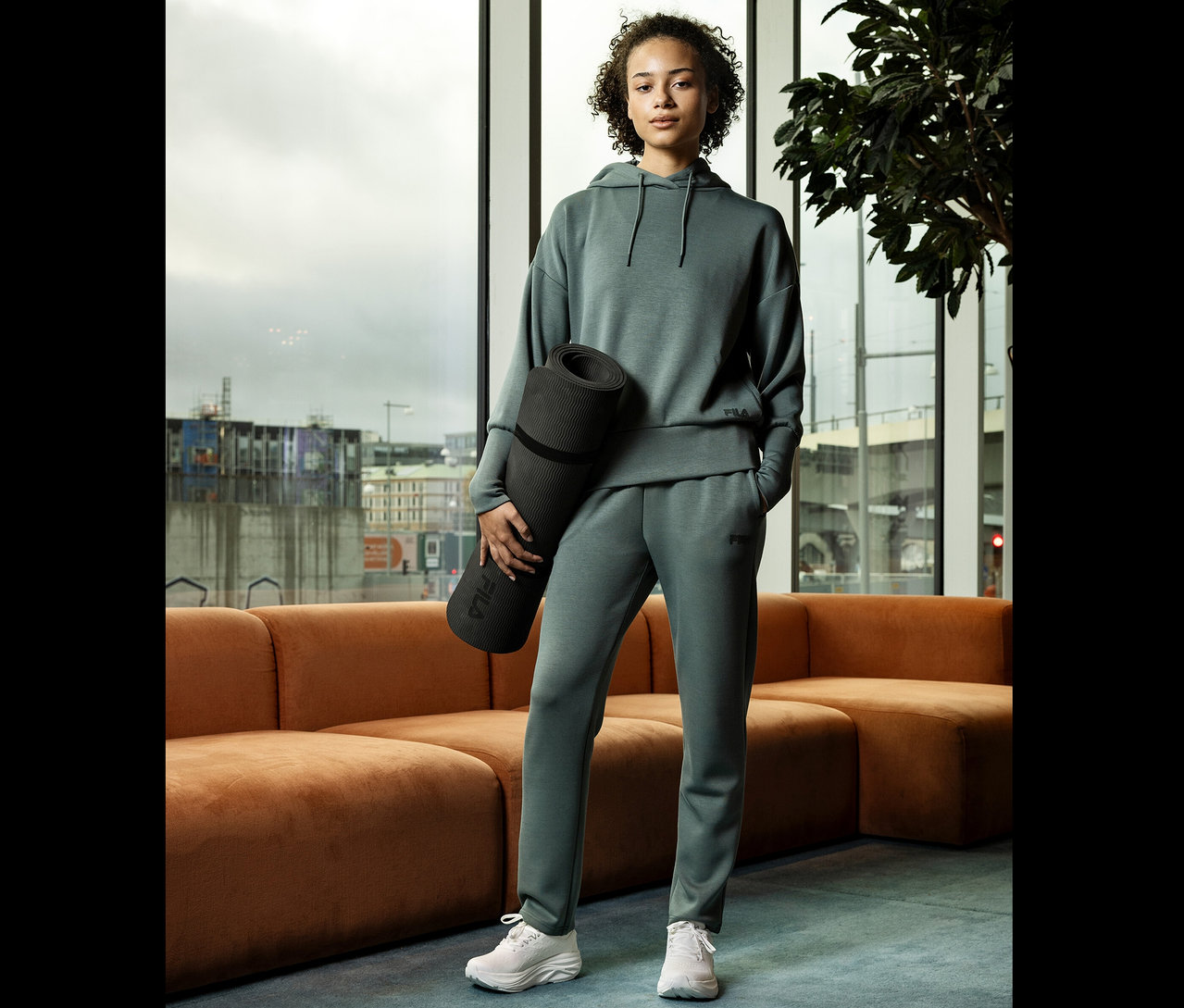 Mladá žena v sivom športovom oblečení drží zrolovanú podložku na cvičenie jogy pred oknom.