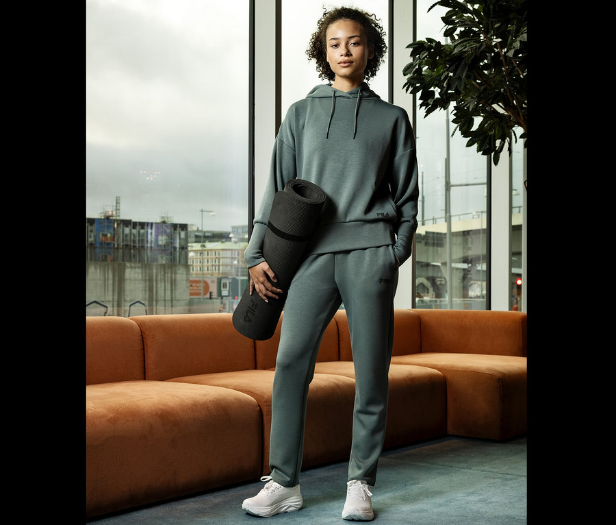 Mladá žena v sivom športovom oblečení drží zrolovanú podložku na cvičenie jogy pred oknom.