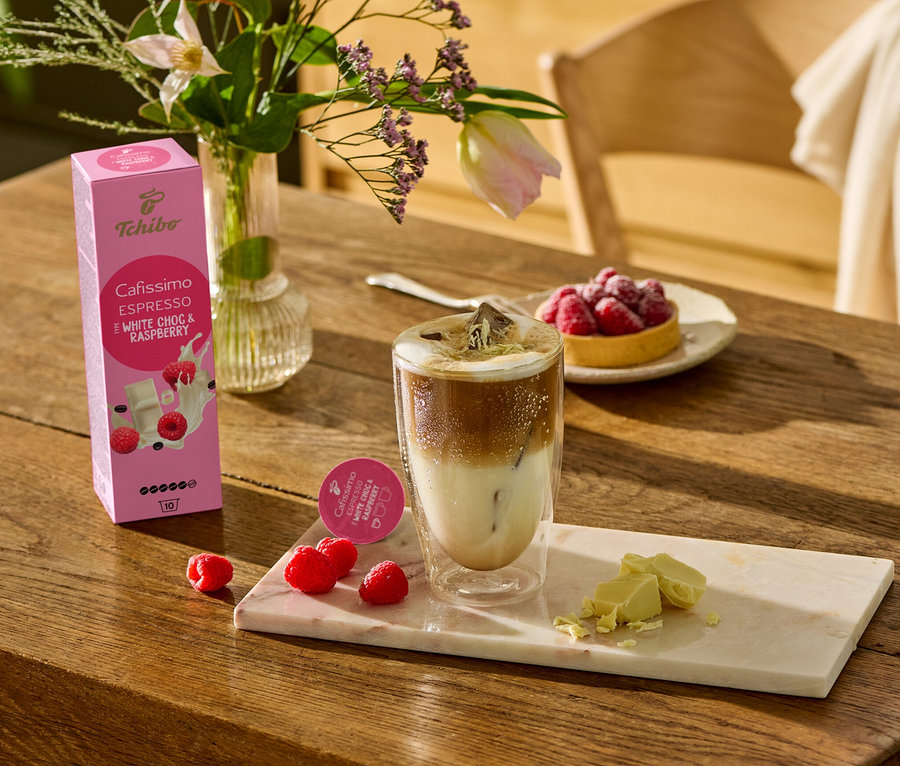 Na drevenom stole stojí pohár s kávou, vedľa neho ležia kúsky bielej čokolády a maliny na mramorovej doske. V pozadí je vidieť balenie Cafissimo Flavoured Espresso - White Choc & Raspberry - 10 kapsúl.