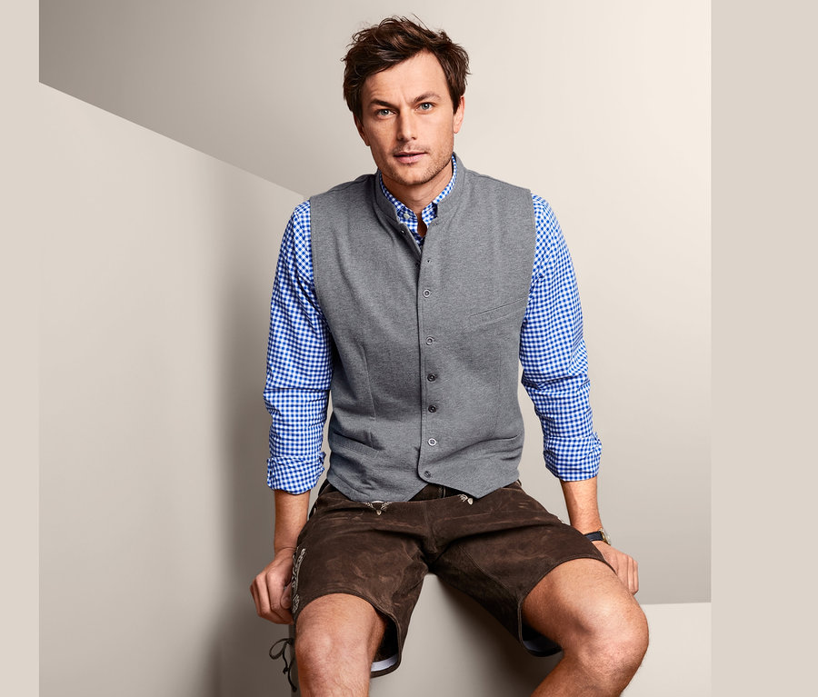 Muž sedí a má na sebe vestu z teplákoviny, košeľu a kožené nohavice.