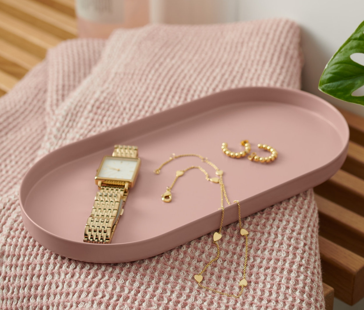 Na drevenej lavici leží kovový podnos hrdzavej farby, na ktorom sú hodinky, náhrdelník a náušnice. Vedľa podnosu leží ružový uterák a fľaša s pumpičkou.