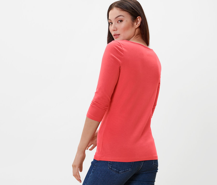 Žena sa pozerá cez plece a má na sebe koralové tričko s trojštvrťovým rukávom a džínsy.