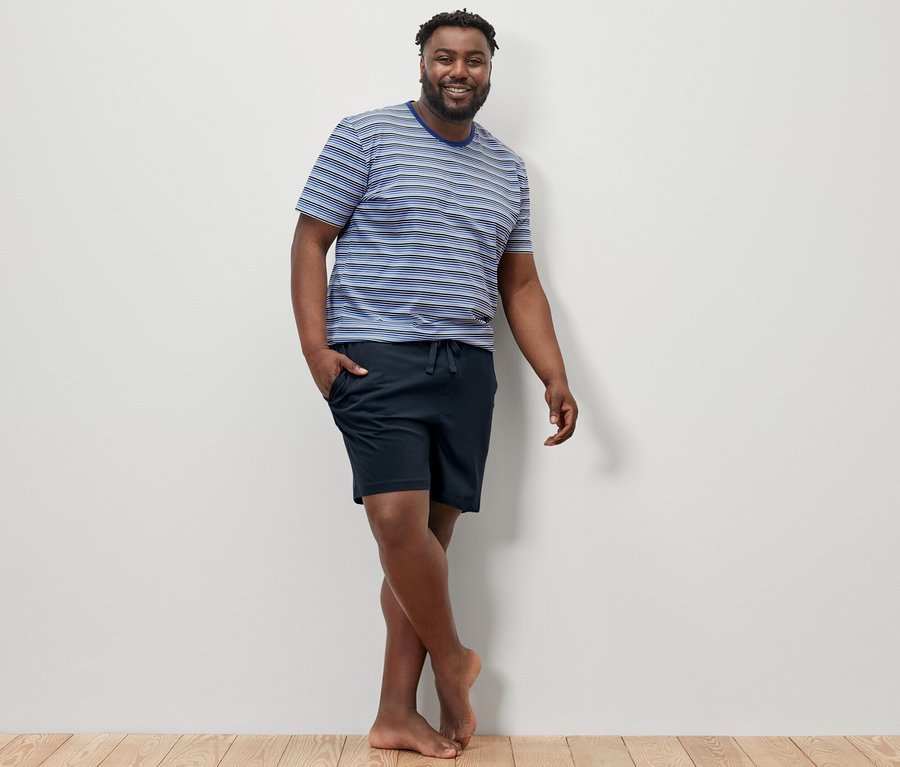 Muž pózuje na drevenej podlahe a má na sebe modro-tmavomodré krátke pyžamo.