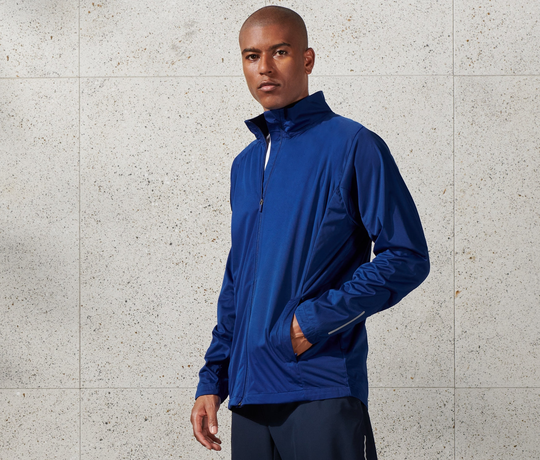 Muž má na sebe modrú vetruvzdornú funkčnú bundu, šortky a biele tenisky.