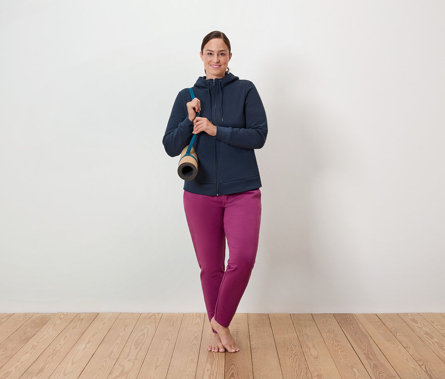 Žena stojí na drevenej podlahe, cez rameno má prehodenú podložku na cvičenie a má na sebe modrú mikinu na zips a fialové nohavice.