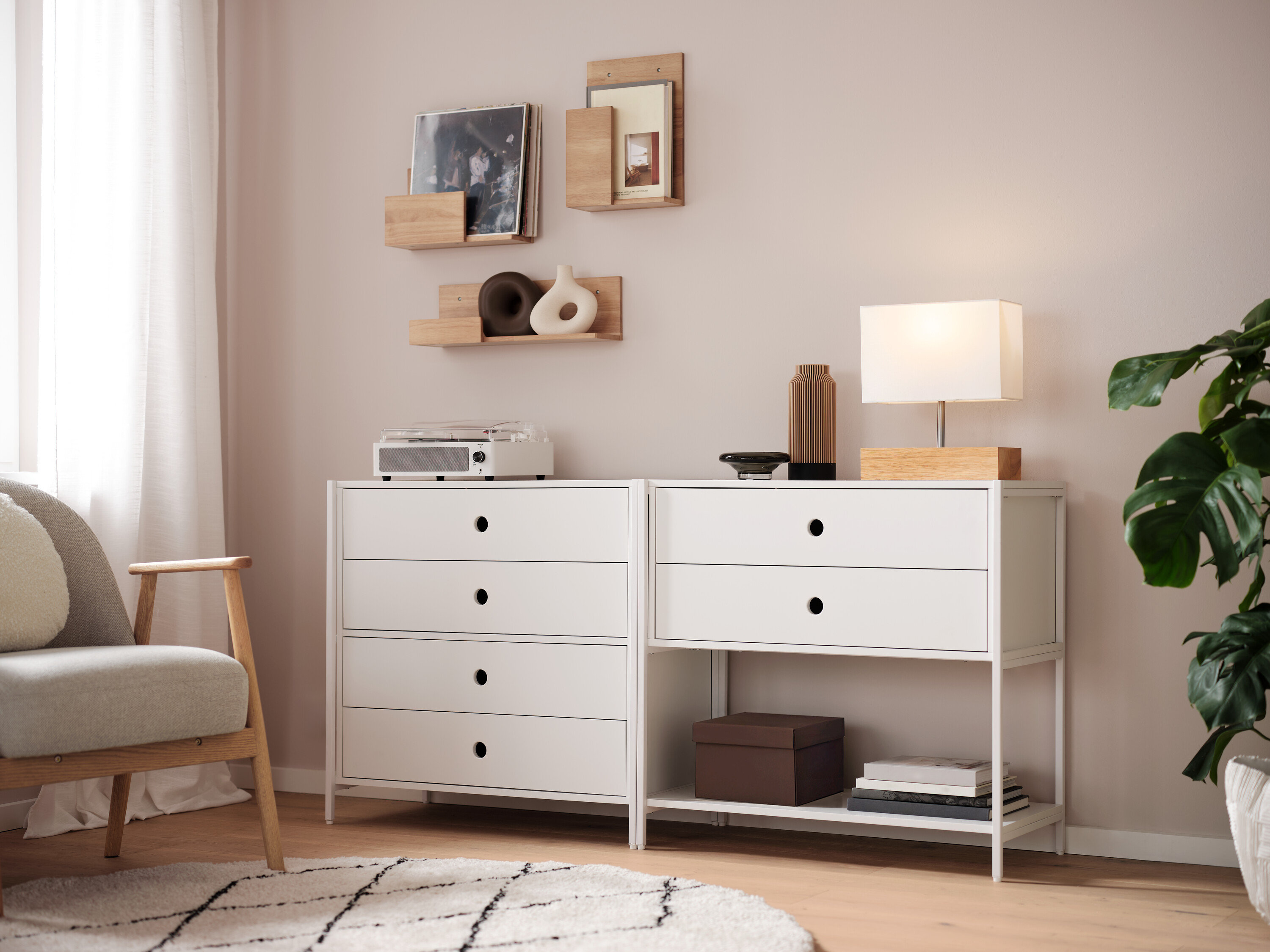 Dve biele komody stoja vedľa seba, na nich je lampa, váza a rádio. Na stene sú police.