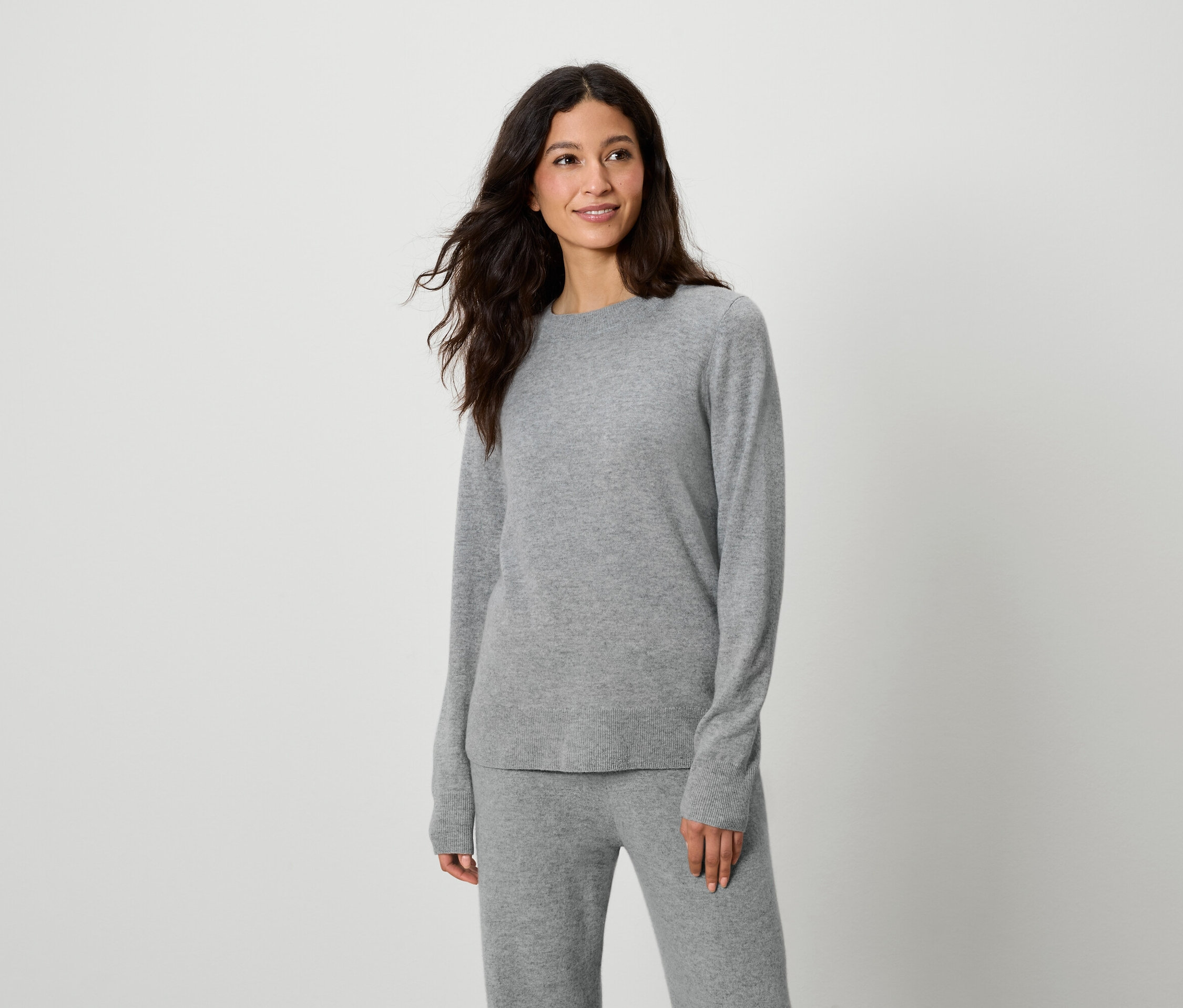 Žena pózuje v sivom svetri a nohaviciach.