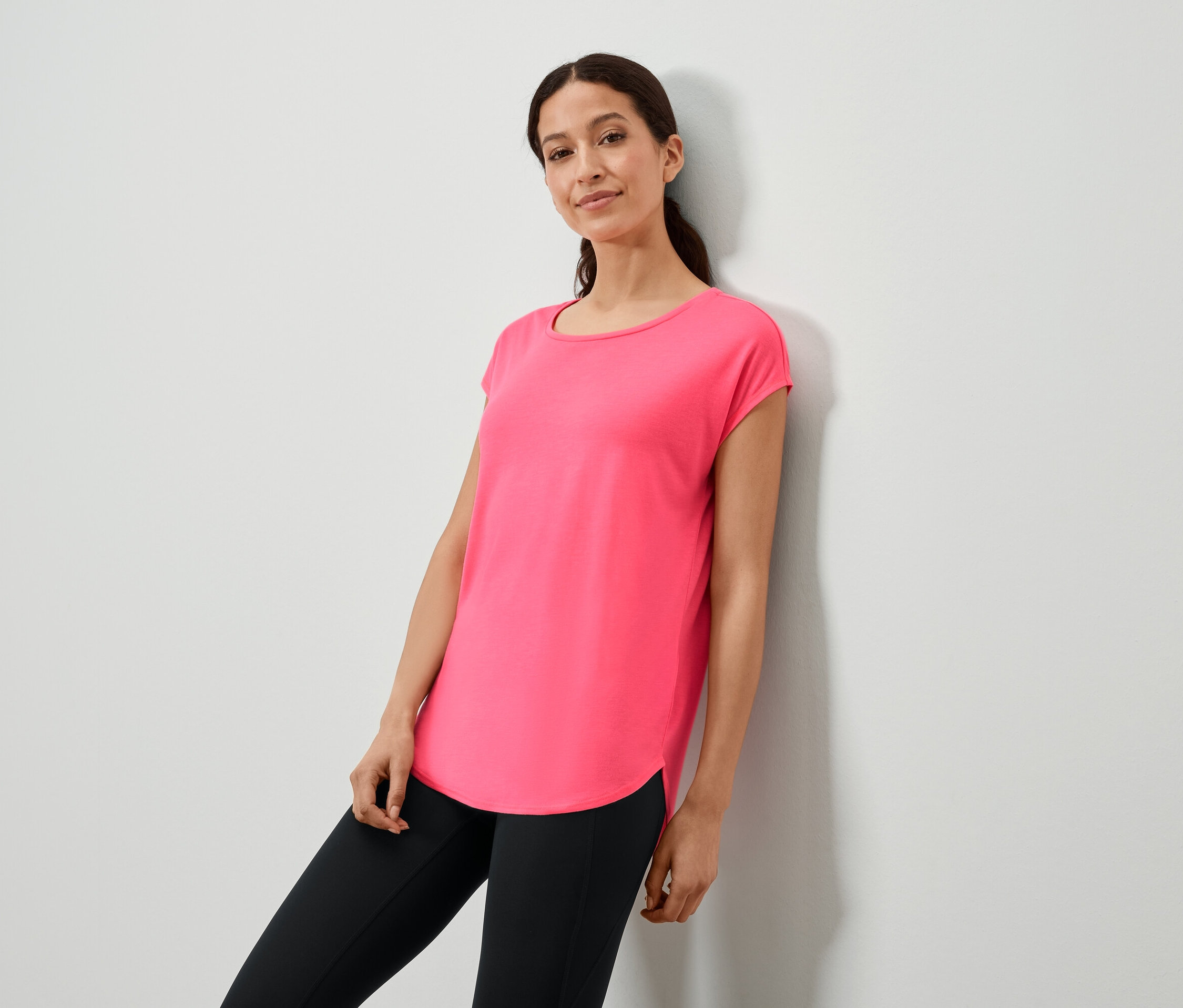 Žena opretá o stenu v ružovom tričku a čiernych legínach.