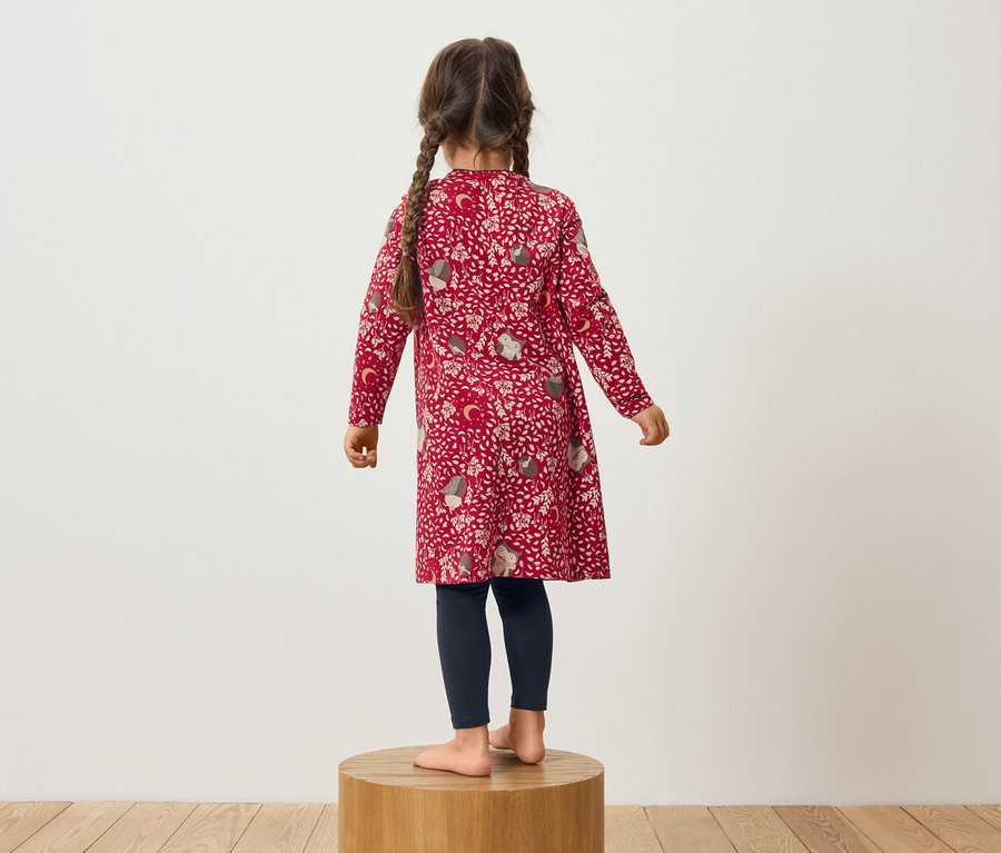 Dievča v červených vzorovaných šatách a tmavomodrých legínach stojí na drevenom podstavci.