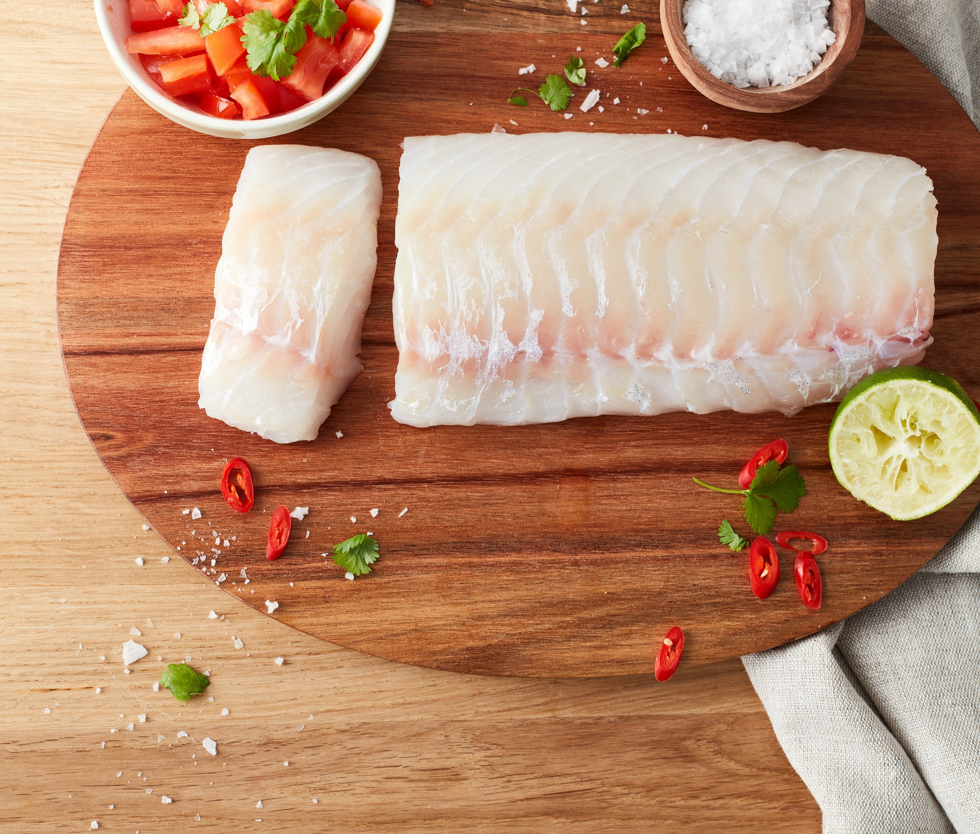 Dva kusy tresky ležia na drevenej doske s nakrájanými paradajkami, soľou, chilli a limetkou.