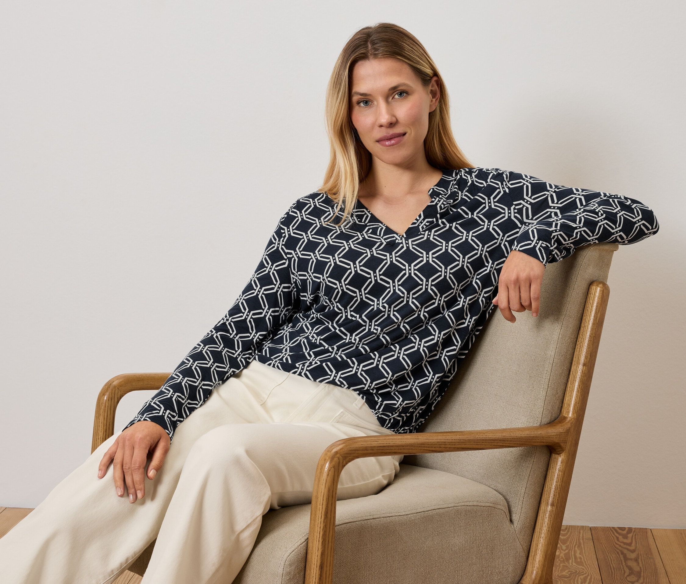 Žena sedí v kresle a má na sebe modro-biely top a krémové nohavice.