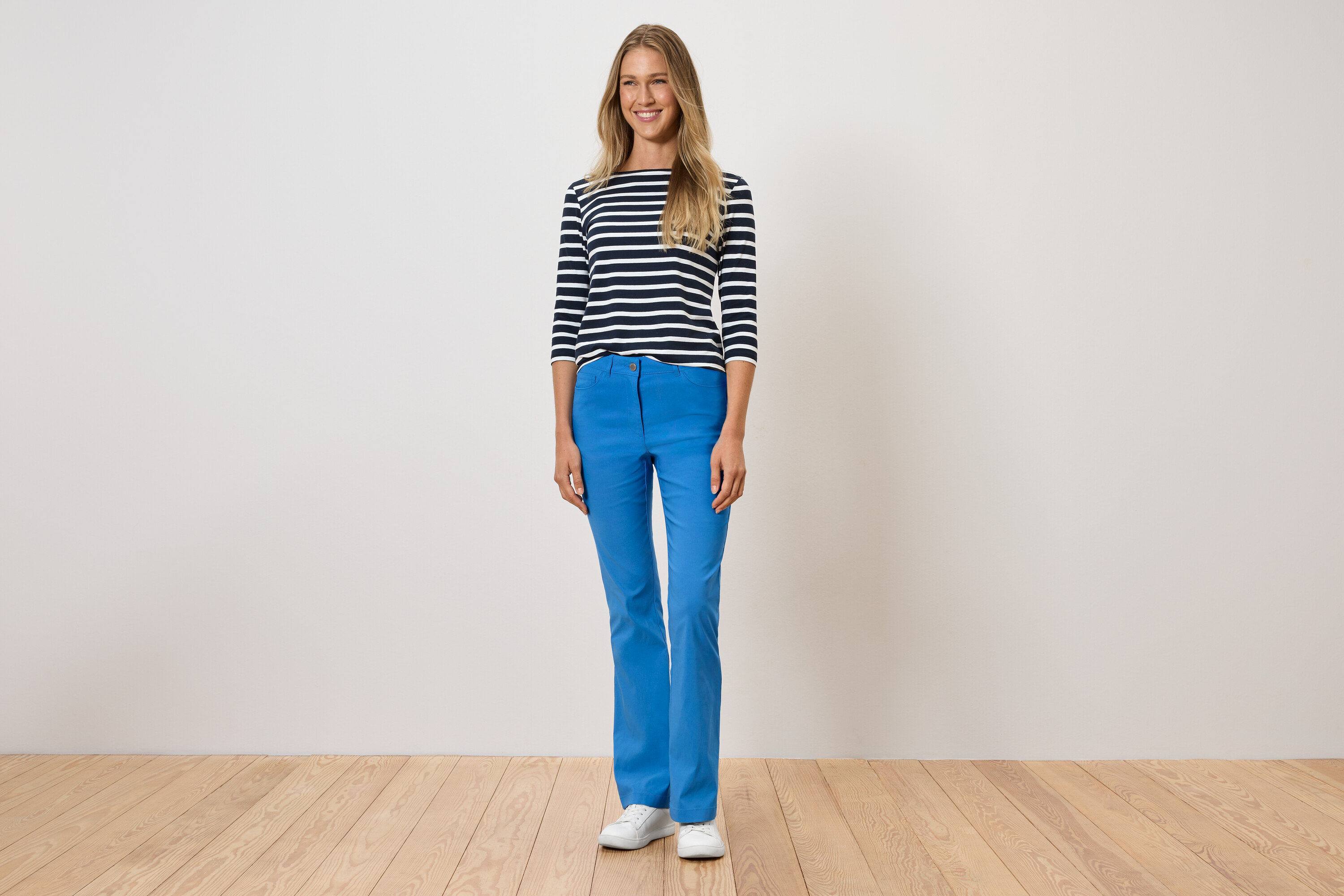 Žena stojí a má na sebe modré nohavice, pruhované tričko a biele topánky.
