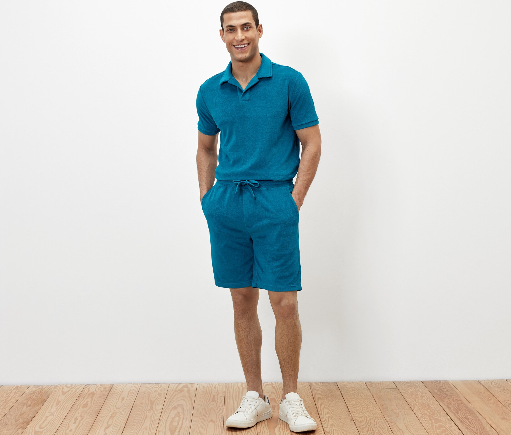 Muž stojí a má ruky vo vreckách froté šortiek petrolejovej farby.