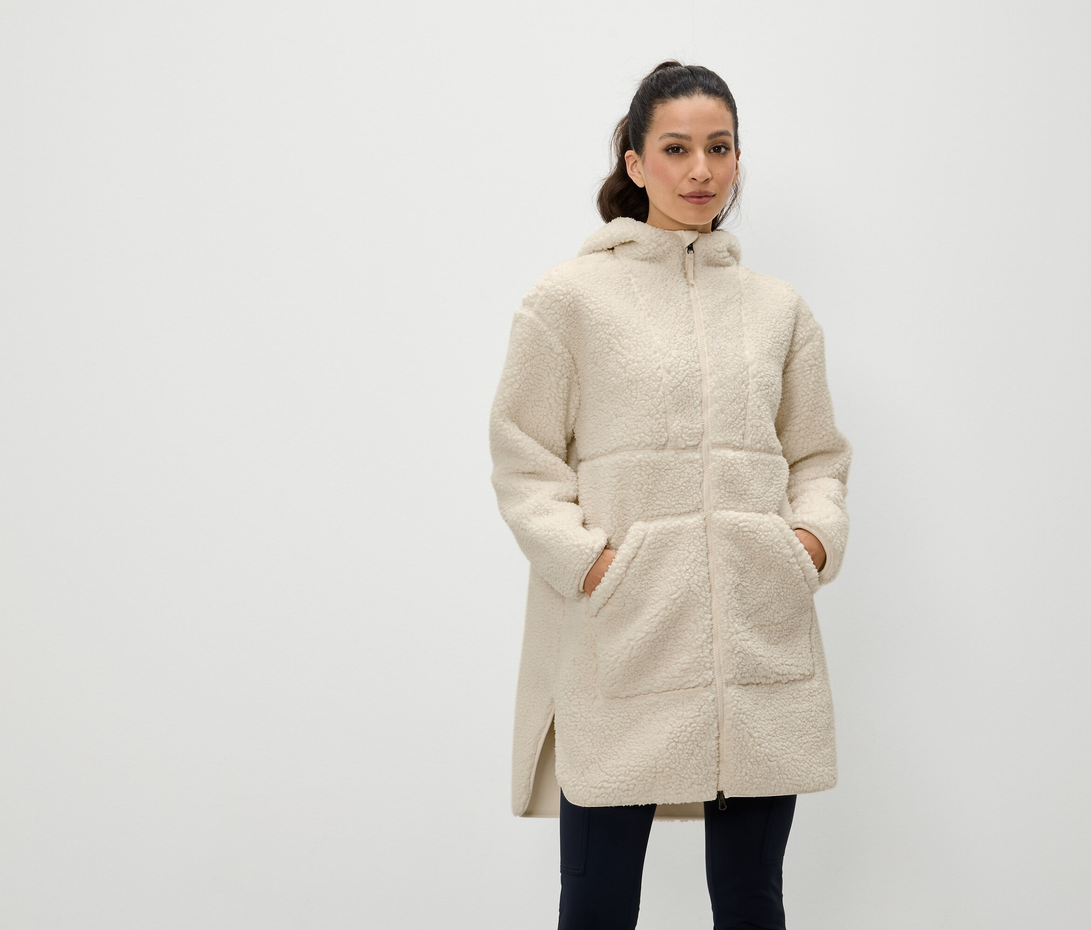 Žena stojí s rukami vo vreckách krémového kabáta s kapucňou na zips.