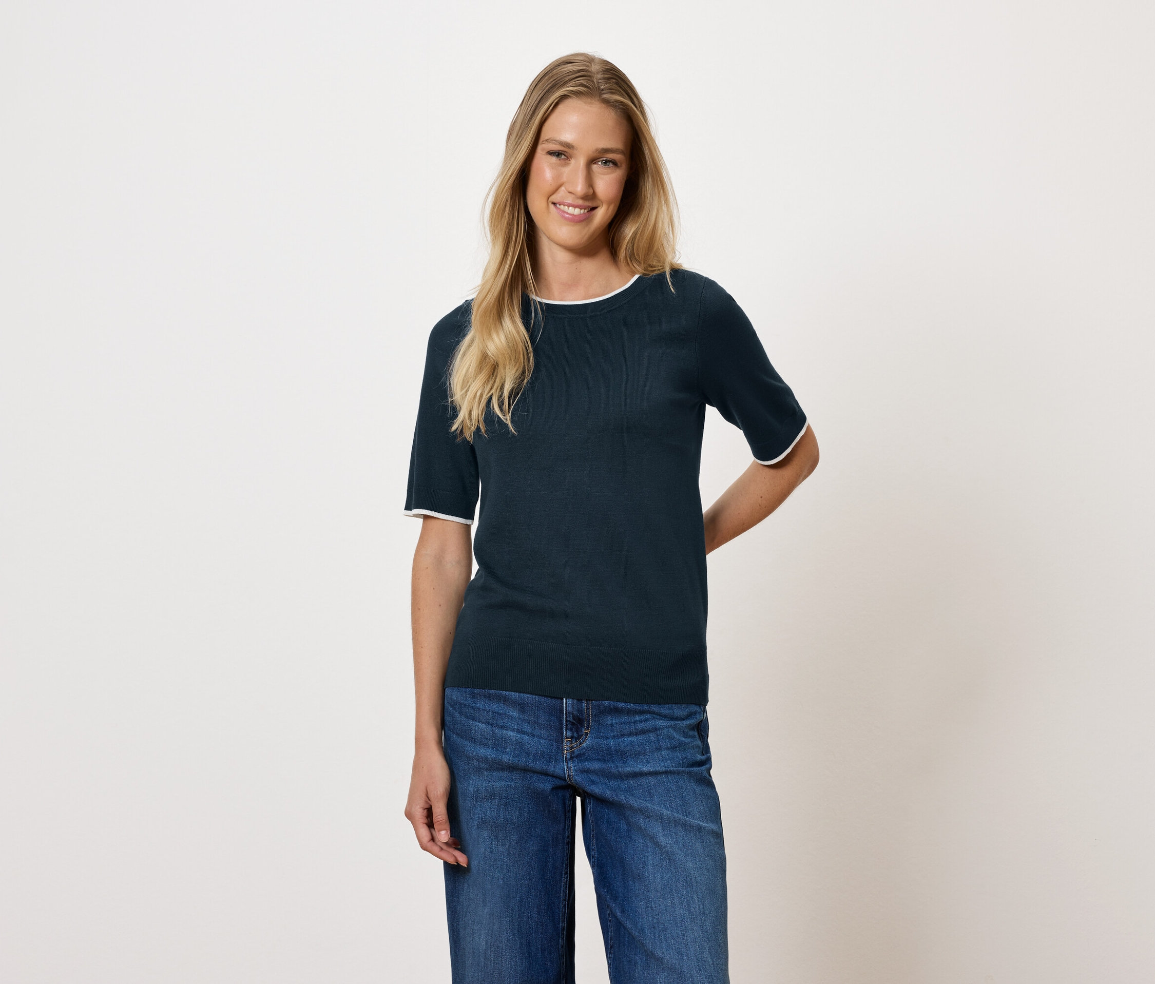 Žena s blond vlasmi má na sebe tmavomodrý top s krátkym rukávom a džínsy.