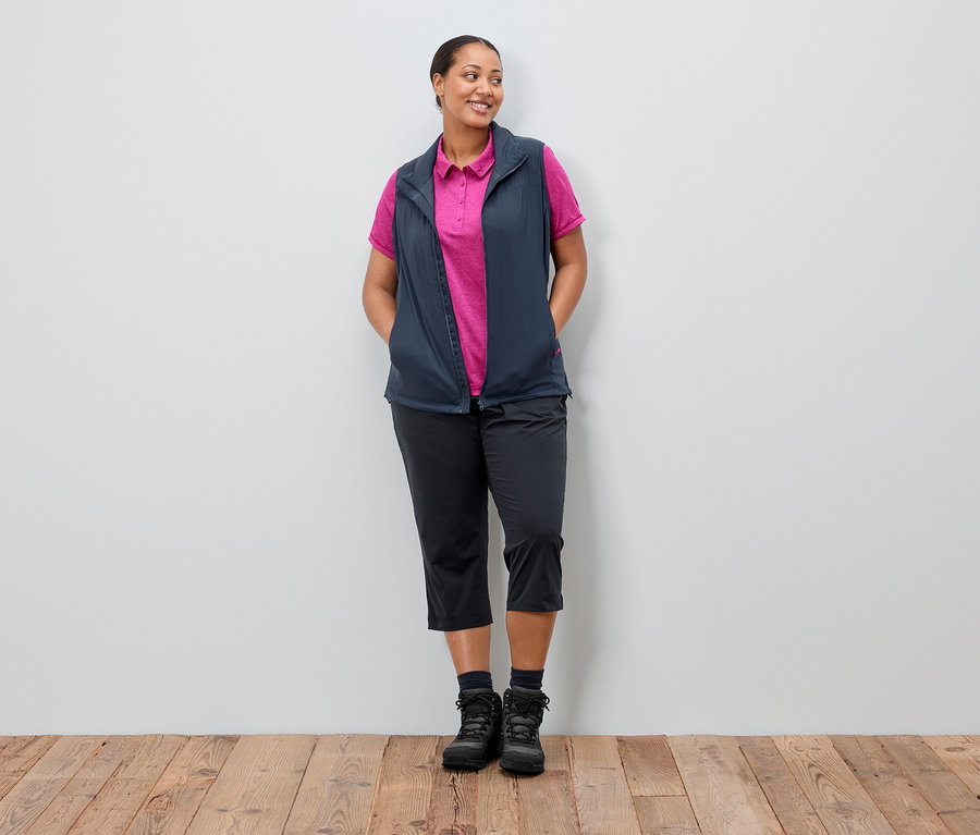Žena stojí s rukami vo vreckách pred stenou. Má na sebe tmavomodrú vestu cez ružové polokošele, čierne nohavice a topánky.