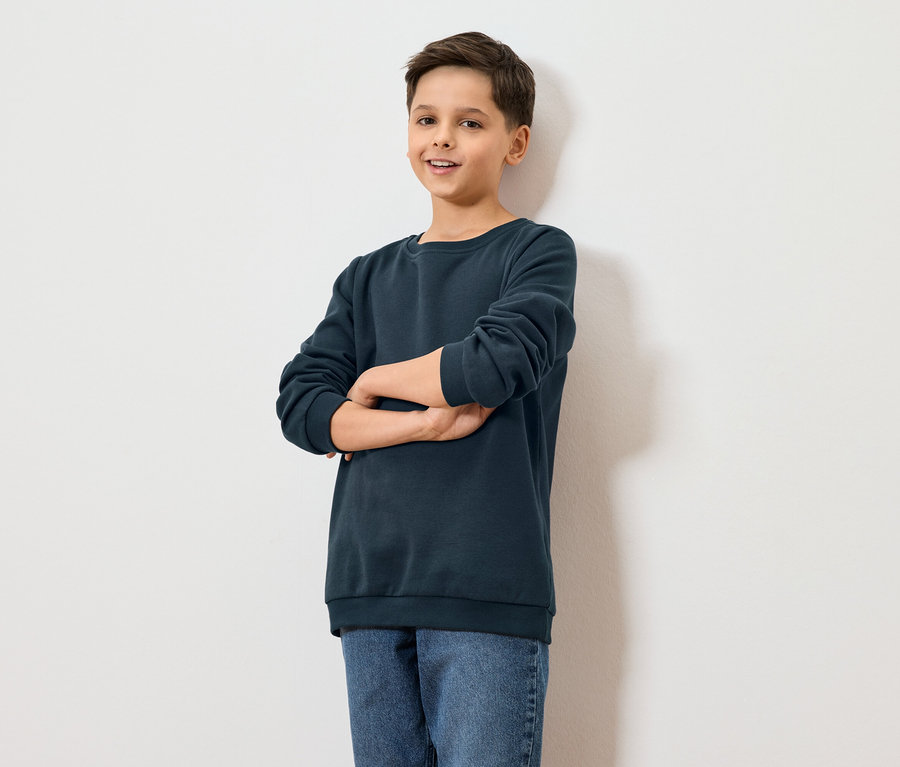 Chlapec stojí s prekríženými rukami pred bielou stenou.