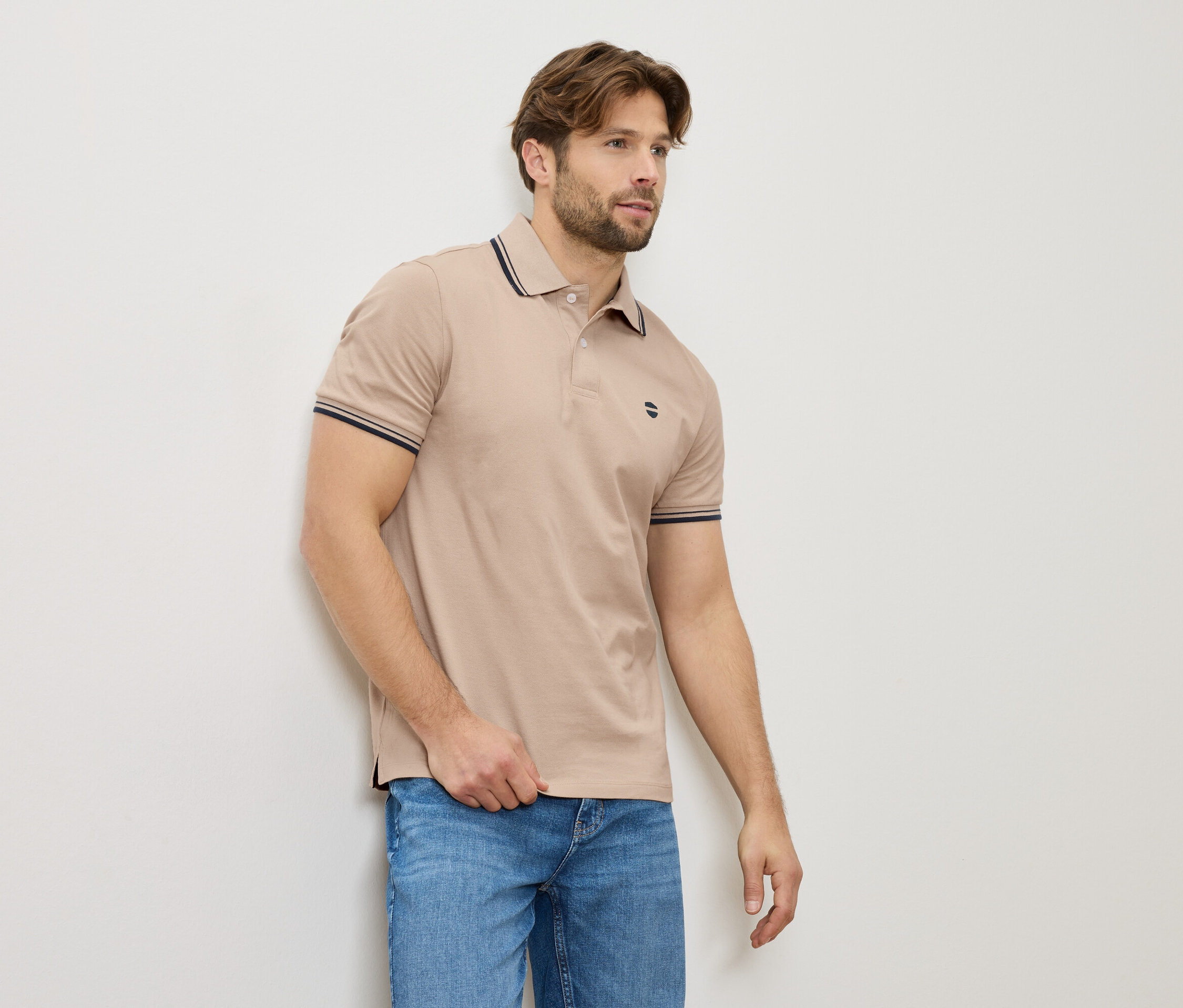 Muž v béžovej polokošeli a džínsoch stojí pri stene.