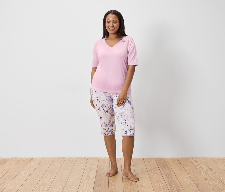 Žena v ružovom tope a kvetovaných nohaviciach stojí naboso na drevenej podlahe pred bielou stenou.