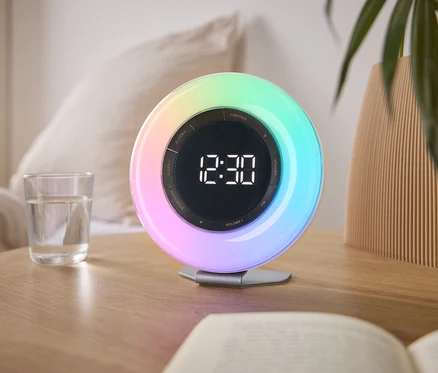 Na nočnom stolíku stojí farebný svetelný budík ukazujúci čas 12:30, vedľa pohára vody a knihy.