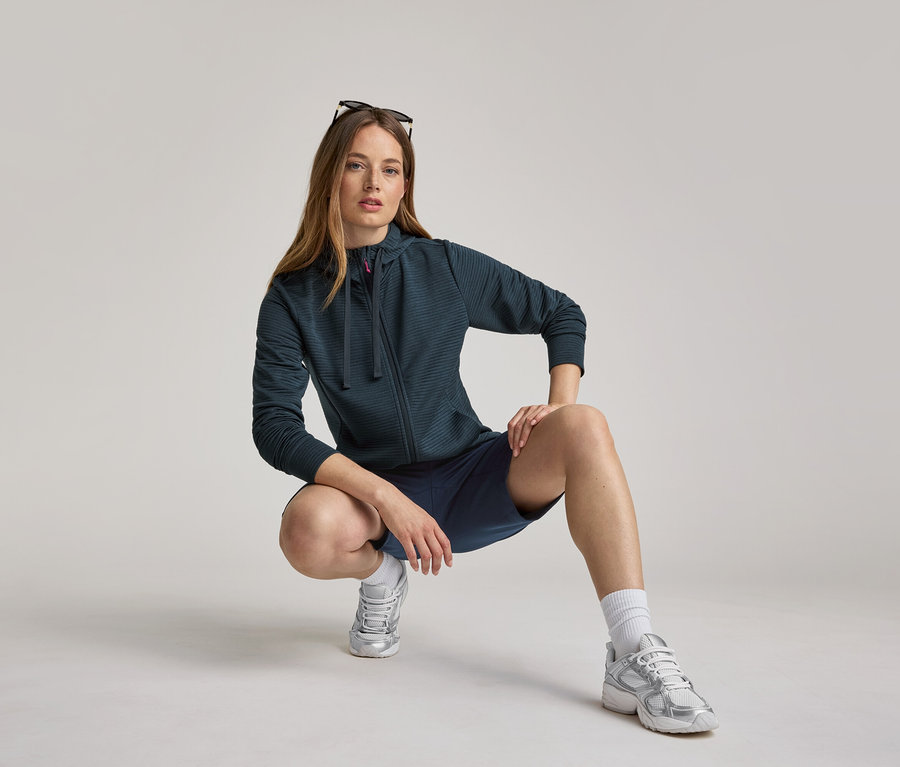 Žena v námorníckej modrej mikine s kapucňou a šortkách čupí. Mikina s kapucňou je námornícka modrá.