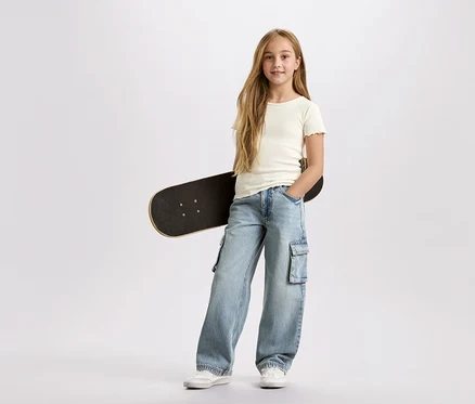Dievča s dlhými blond vlasmi, s čiernym skateboardom pod pazuchou, má na sebe svetlomodré cargo nohavice a biele tričko na bielom pozadí.