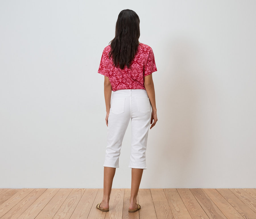 Žena s tmavými vlasmi stojí chrbtom k fotoaparátu, má na sebe červený vzorovaný top, biele trojštvrťové nohavice a zlaté sandále.