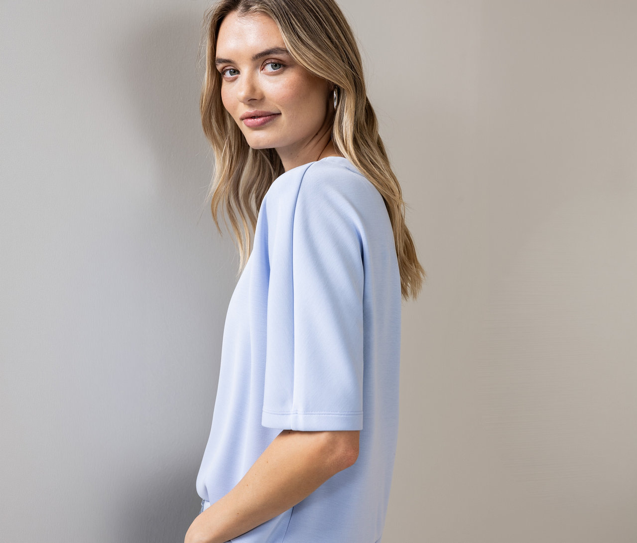 Žena sa pozerá cez plece, má na sebe svetlomodré tričko Street One s krátkymi rukávmi a strieborné kruhové náušnice.