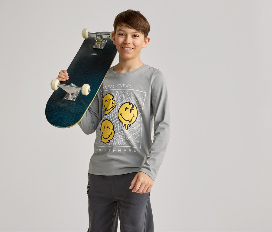 Usmievavý chlapec drží skateboard na ramene, má na sebe sivé tričko s dlhým rukávom so žltými motívmi smajlíkov a sivé nohavice.