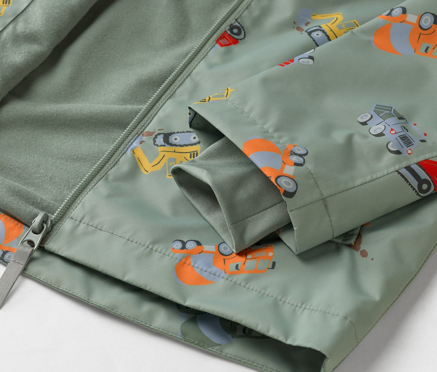 Detailný záber na olivovozelenú bundu so vzorom stavebných vozidiel, čiastočne rozopnutú na zips.
