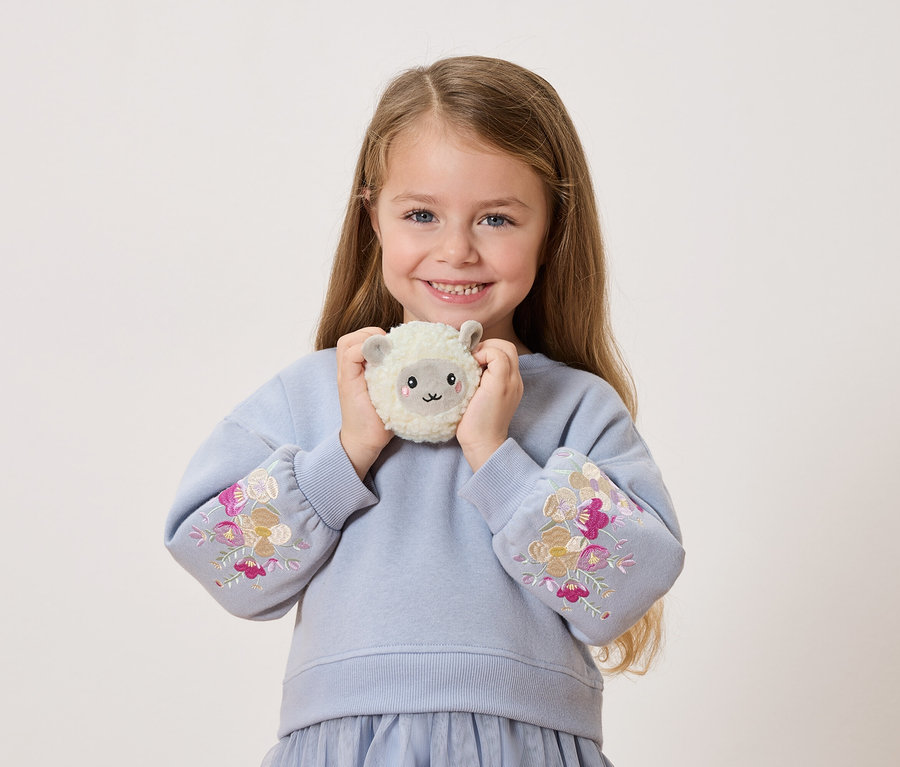 Usmievavé dievčatko drží pred sebou plyšovú ovečku. Má na sebe modrý sveter s kvetinovými výšivkami na rukávoch.