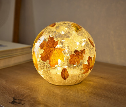 Guľatá lampa s prasknutou sklenenou textúrou, ozdobená jesennými listami, teplo svieti na drevenom povrchu. Malé svetielka vo vnútri osvetľujú listy.