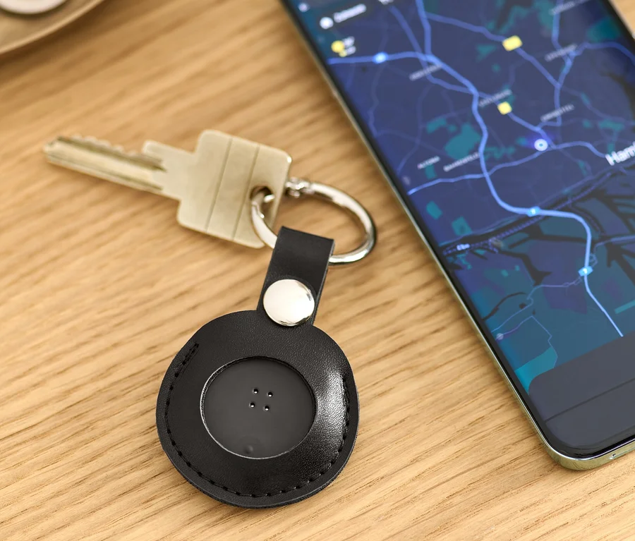 Na stole ležia kľúče s čiernym Smart Tracker s príveskom a mobilný telefón s mapou.