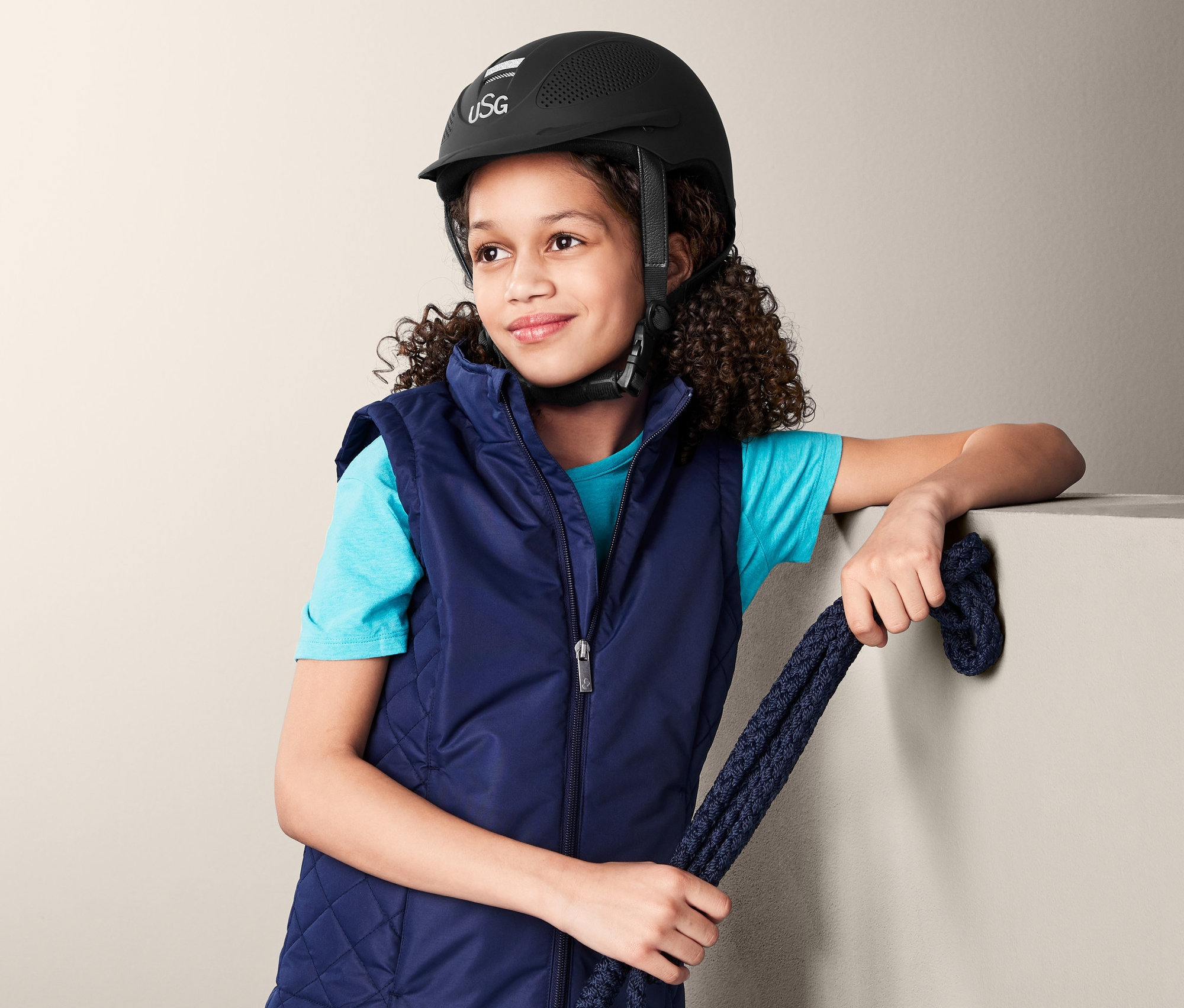 Dievča s hnedými kučeravými vlasmi má na sebe modrú jazdeckú vestu a čiernu detskú jazdeckú prilbu USG a drží modré lano.