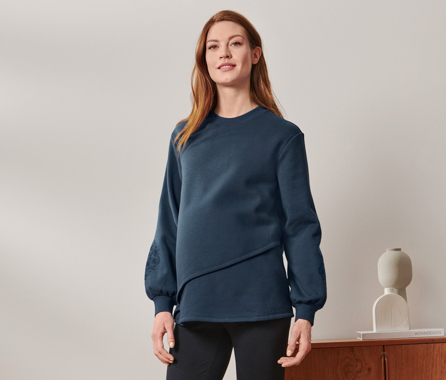 Tehotná žena stojí a má na sebe tehotenskú a dojčiacu mikinu, čierne nohavice a tenisky.