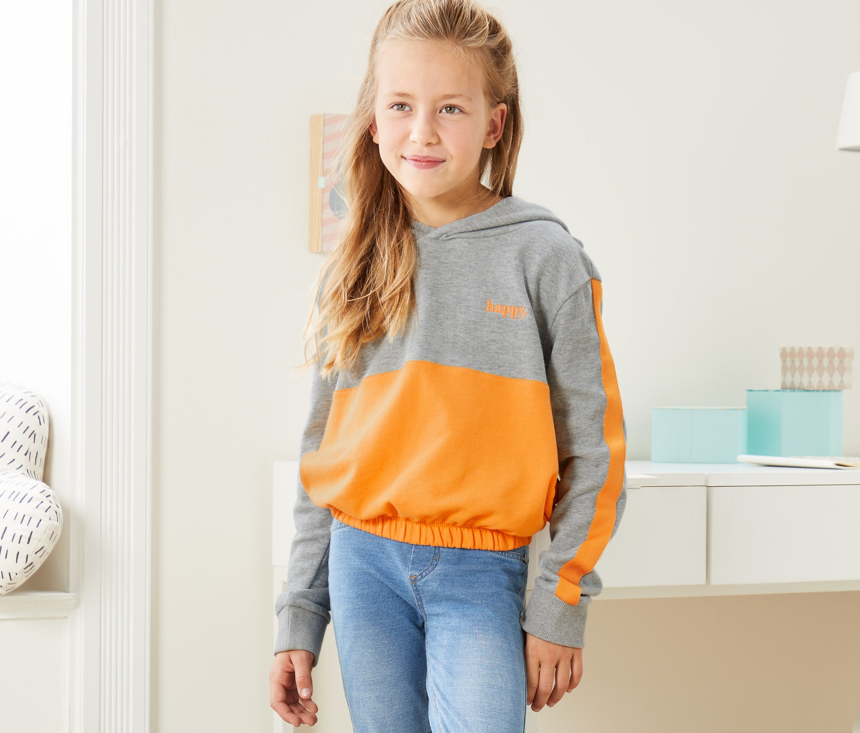Dievča stojí a má na sebe sivo-žltú melírovanú mikinu s kapucňou v skrátenej dĺžke, džínsy a tenisky.