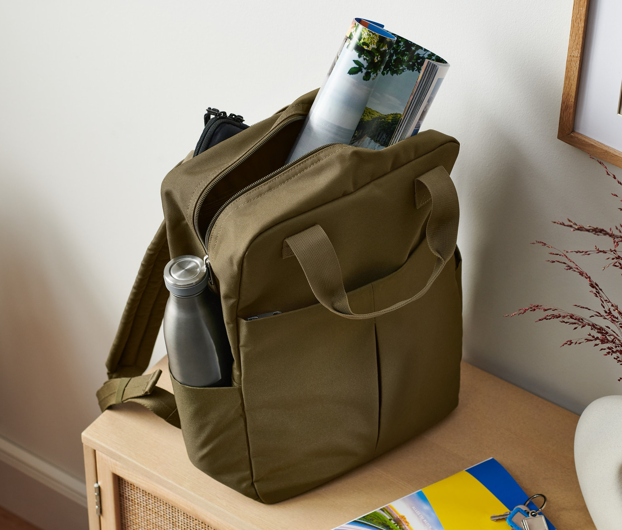 Khaki bezpečnostný ruksak s fľašou na vodu v bočnom vrecku a časopisom vnútri.