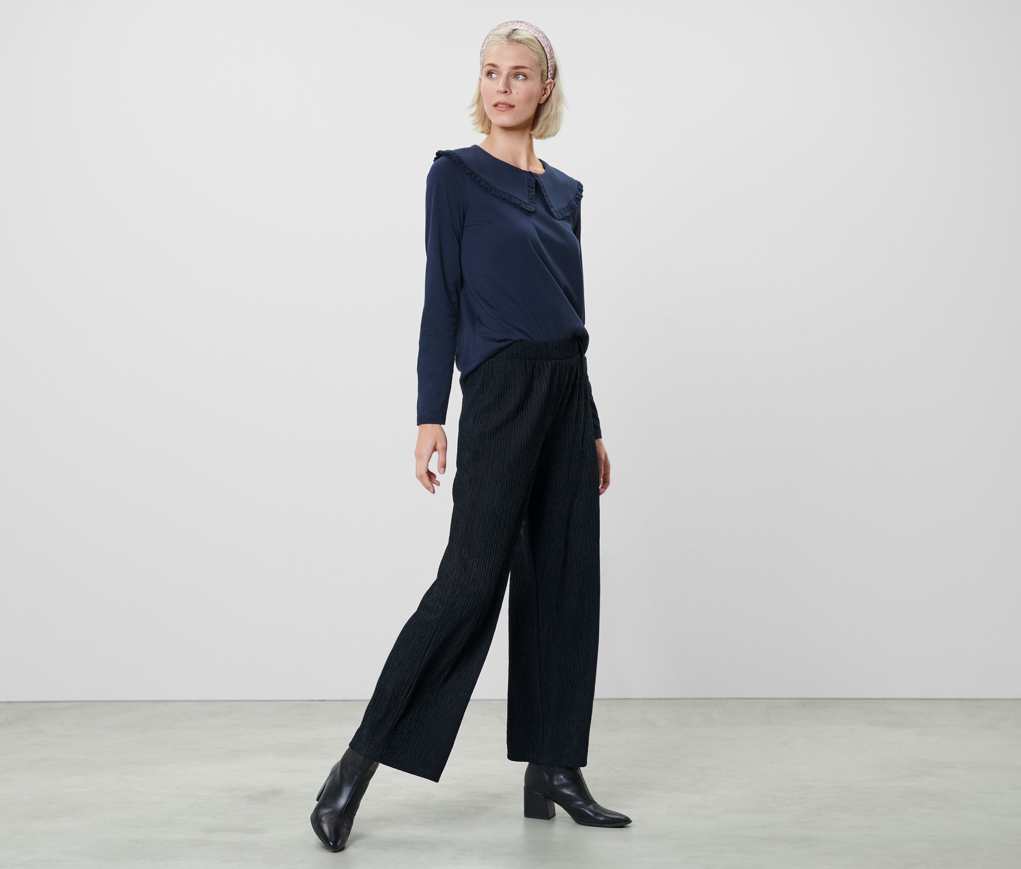 Žena pózuje v tmavomodrej blúzke a tmavomodrých plisovaných nohaviciach.